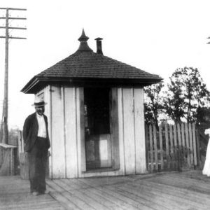 man in straw hat outside small building with pointed roof standing alongside railroad tracks