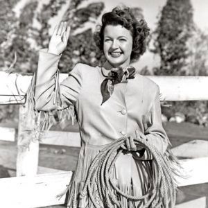 White woman in western clothes holding a rope in left hand and raising her right in greeting