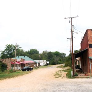 Dirt road with brick and stone storefronts on the left side and multistory buildings with covered porches on the right side